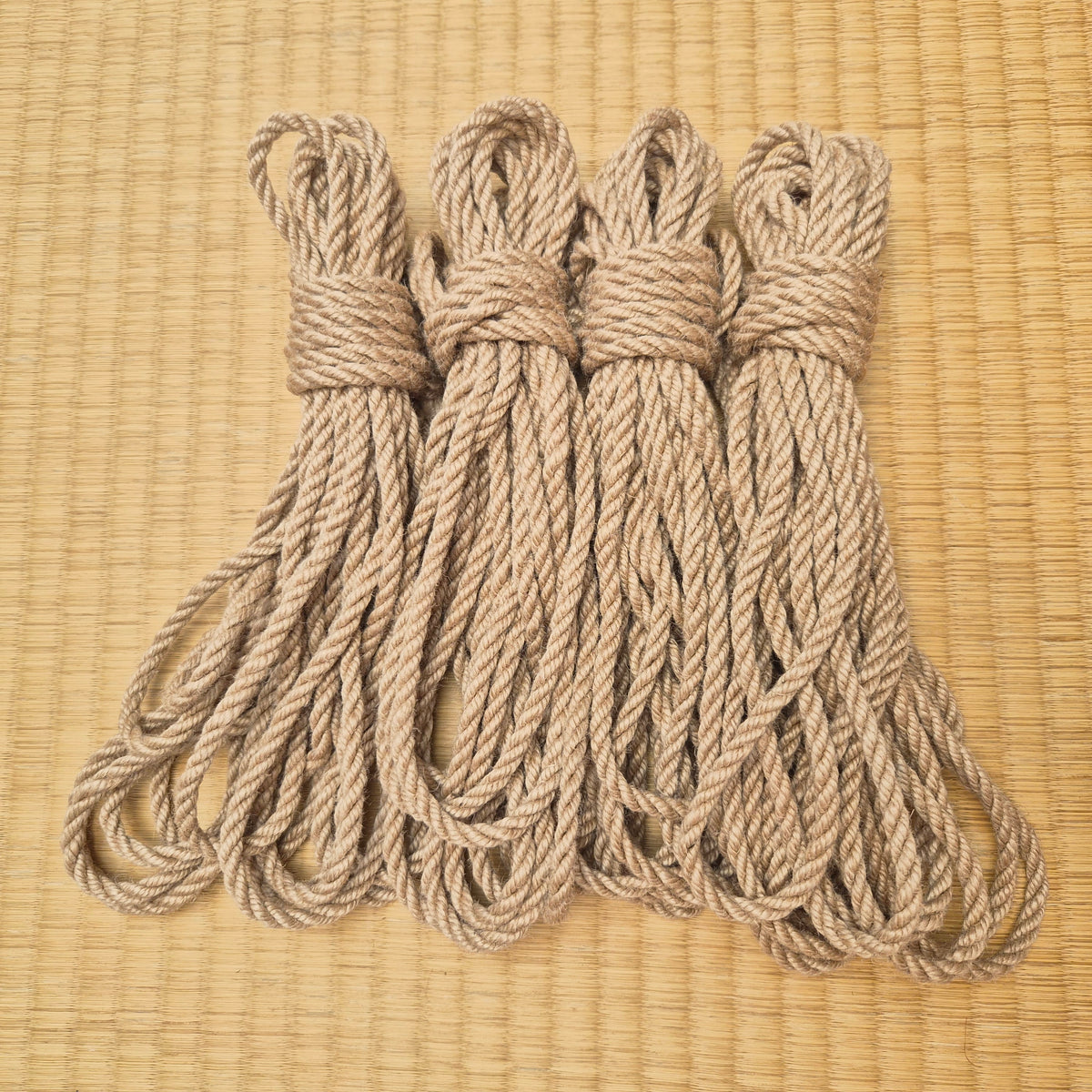 Four coiled natural fiber kinbaku ropes in a light tan color arranged side by side on a woven straw mat background.