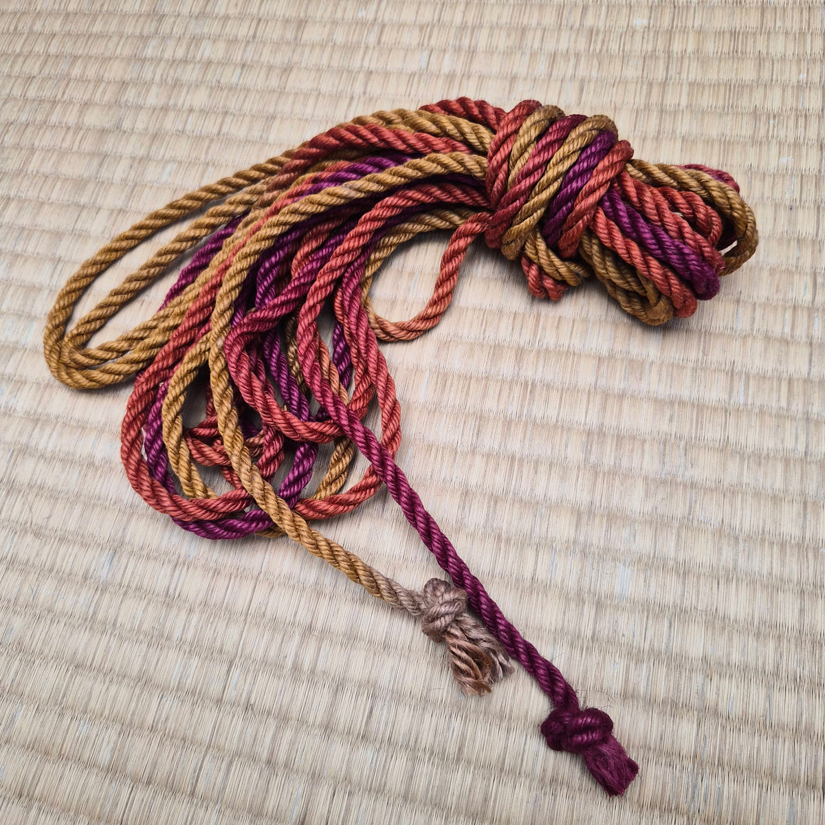 A coiled bundle of shibari rope in mixed warm tones, including tan, rust red, and deep burgundy, placed on a woven straw mat background.