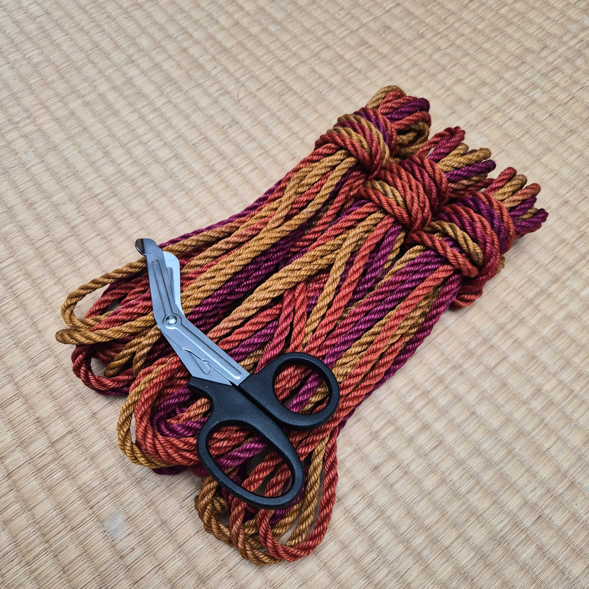 A bundle of twisted yarn or cord in mixed warm tones of burgundy, rust orange, and gold, with a pair of black-handled scissors resting on top, photographed on a woven beige mat.