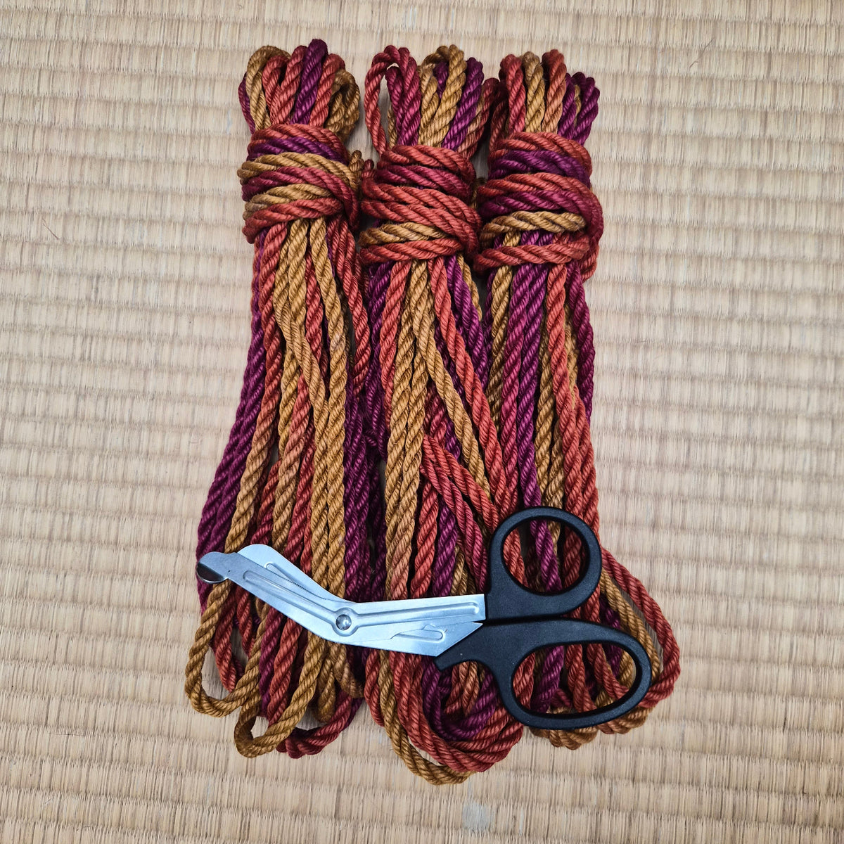 Three bundled strands of twisted jute rope in a variegated pattern of gold, rust orange, and deep burgundy, arranged on a woven straw mat with a pair of black-handled scissors in the foreground.