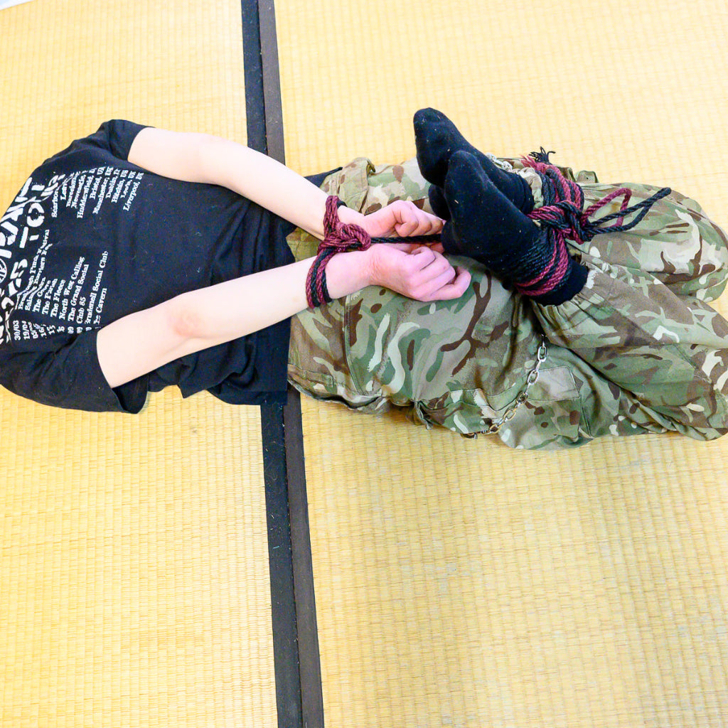 Person in camouflage pants and black shirt sitting on a yellow mat with hands tied behind their back.
