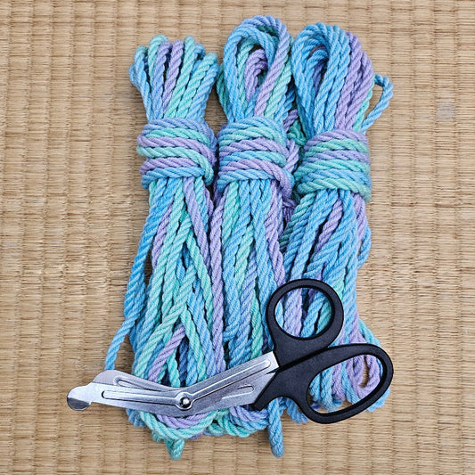 Three bundled skeins of variegated yarn in light blue, teal, and lavender-purple, arranged on a tan woven mat with a pair of silver and black scissors in front.