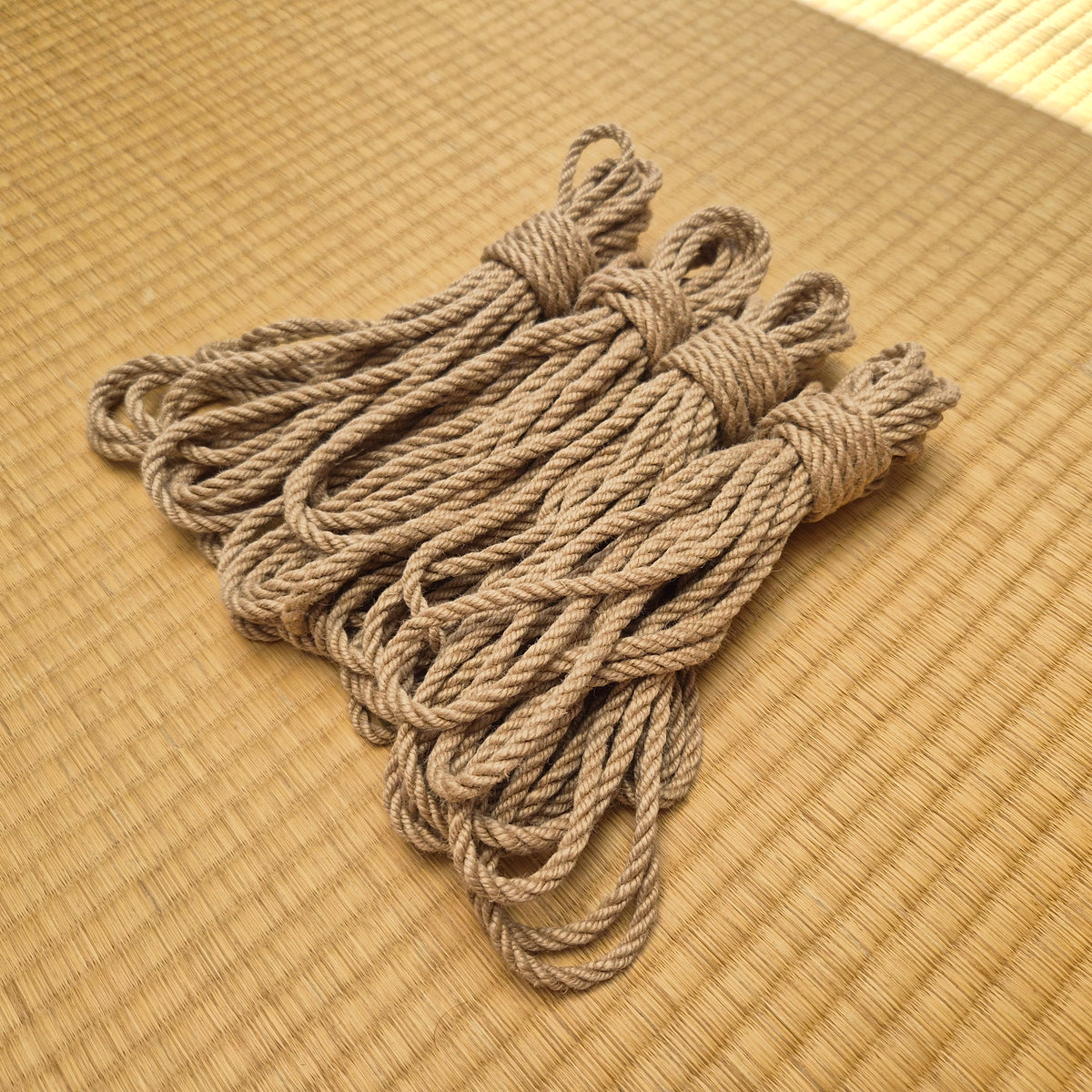 A bundle of 4 natural tan kinbaku ropes tied in hanks, arranged on a woven straw mat background.