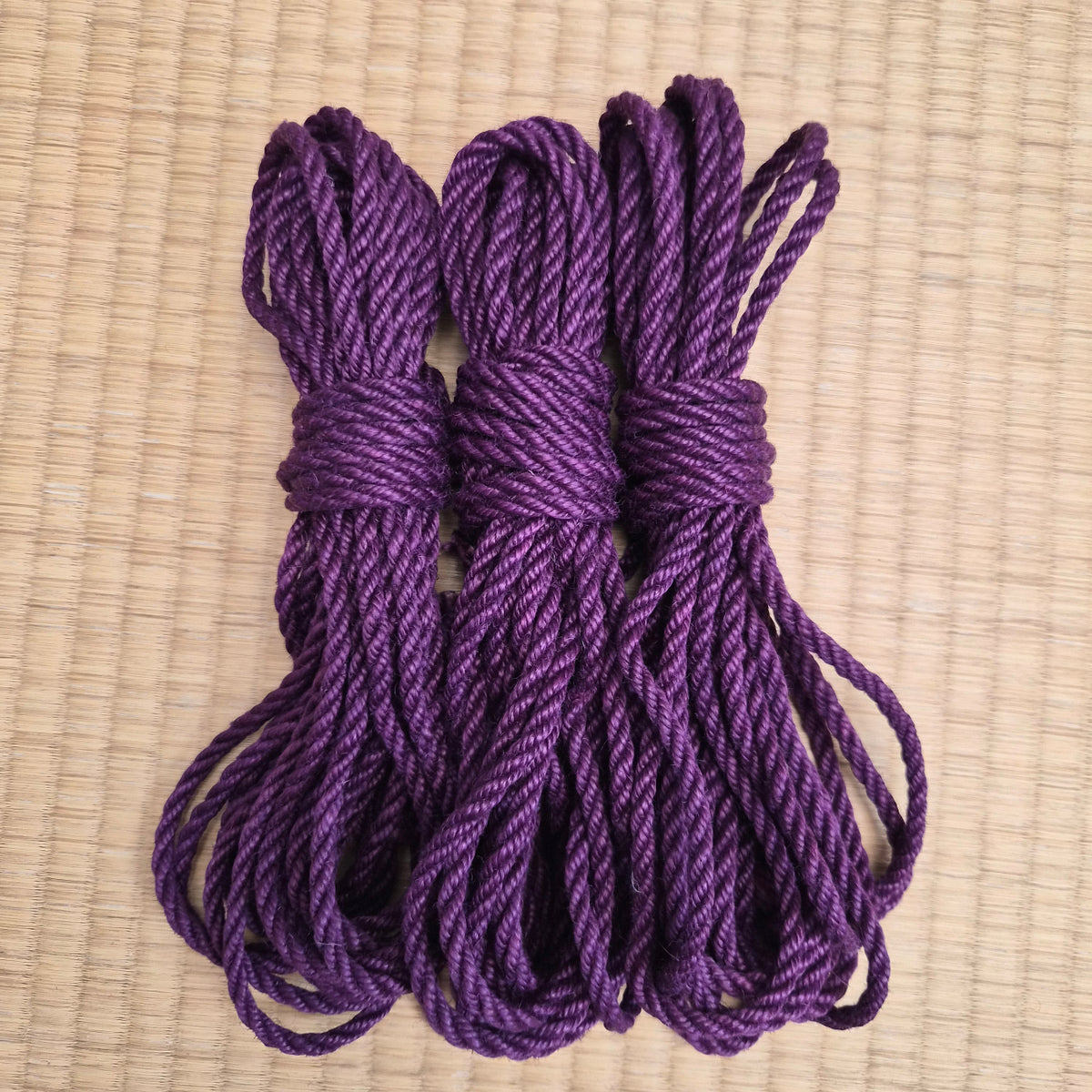 Three coiled bundles of purple twisted rope or cord tied together, displayed on a light beige woven mat background.